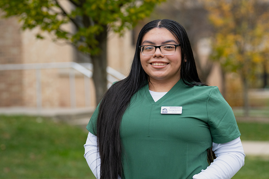 A medium-length portrait of a smiling woman with long, straight dark hair and glasses. She is wearing green medical scrubs over a white long-sleeved shirt and has a professional name tag pinned to her chest. She is standing outdoors with a green lawn, trees, and a brick building in the soft-focus background.
