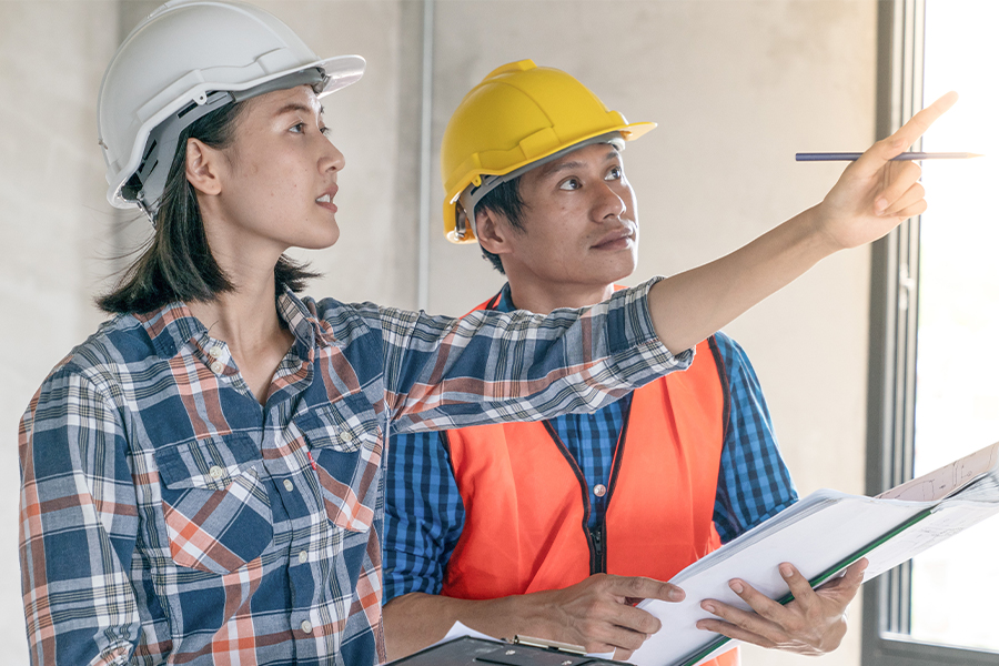 Two construction inspectors wearing hard hats and safety vests review a clipboard together inside a building, with one pointing toward a wall or ceiling feature.