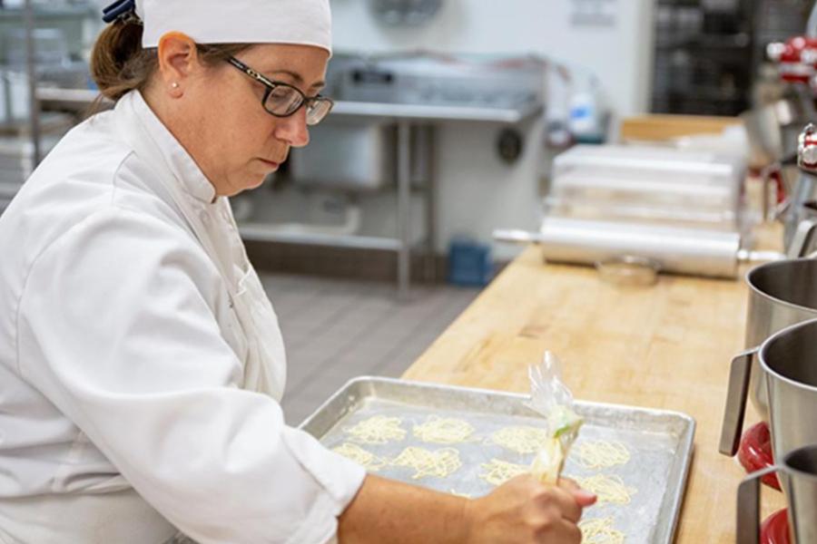 A female chef in a white uniform and hat carefully uses a piping bag to create intricate patterns on a baking sheet in a professional kitchen setting.
