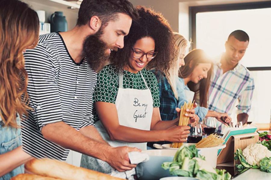 A diverse group of smiling adults cooking a meal together in a brightly lit kitchen.