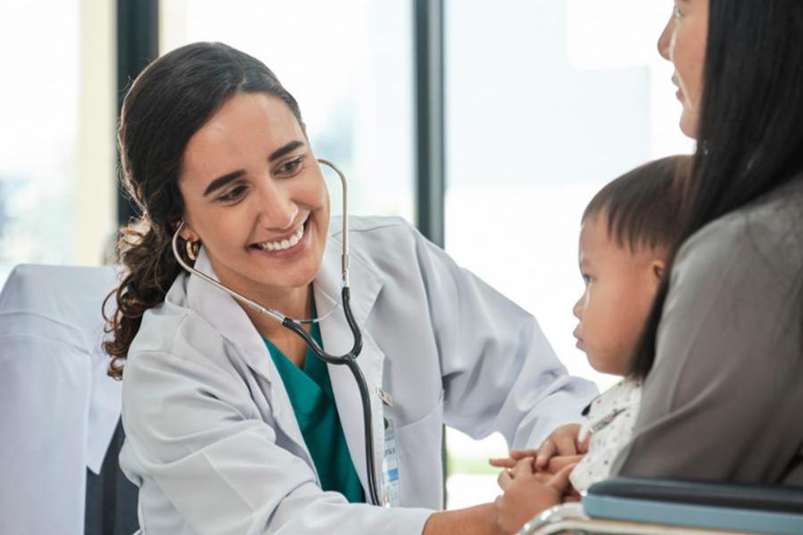 Smiling medical professional in a white coat holding a stethoscope while examining a toddler seated on a parent's lap in an exam room.